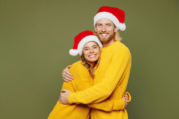 Side view merry young couple two friends man woman wear sweater Santa hat posing hug cuddle embrace look camera isolated on plain green background Happy New Year celebration Christmas holiday concept