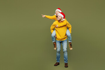 Full body merry young couple two friend man woman in sweater Santa hat posing giving piggyback ride...