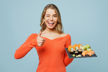 Young smiling happy satisfied woman wear orange casual clothes show thumb up gesture wink hold eat...