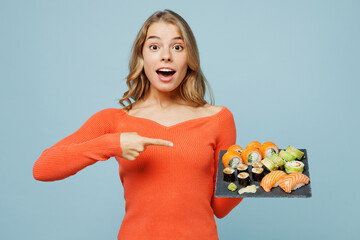 Young surprised shocked fun woman wears orange casual clothes hold eat raw fresh sushi roll served on black plate point index finger on Japanese food isolated on plain blue background studio portrait.