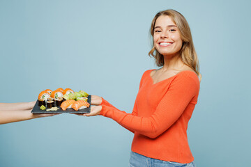 Side profile view young woman wearing orange casual clothes look camera holding eat taking receive raw sushi roll served on black plate Japanese food isolated on plain blue background studio portrait.