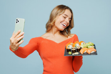 Young happy woman wear orange casual clothes doing selfie shot on mobile cell phone hold eat raw fresh sushi roll served on black plate Japanese food isolated on plain blue background studio portrait.