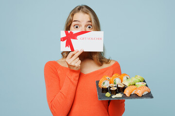Young woman in orange casual clothes hold cover mouth store gift certificate coupon voucher card eat raw fresh sushi roll served on black plate Japanese food isolated on plain blue background studio