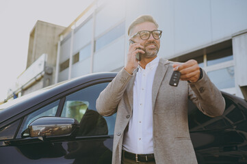Adult happy man customer male buyer client wear white shirt classic grey suit standing near black electric car while charging talk speak on mobile cell phone hold give keys outside. Business concept. - obrazy, fototapety, plakaty