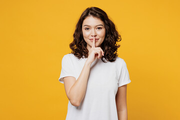 Young secret smiling Caucasian woman she wearing white blank t-shirt casual clothes saying hush be quiet with finger on lips shhh gesture isolated on plain yellow orange background. Lifestyle concept.