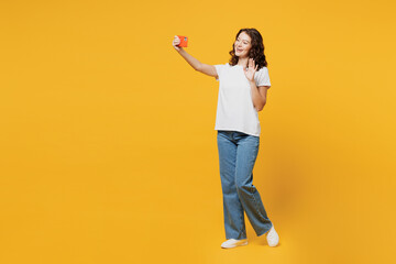 Full body young woman wear white blank t-shirt casual clothes do selfie shot on mobile cell phone post photo on social network waving hand isolated on plain yellow orange background studio portrait