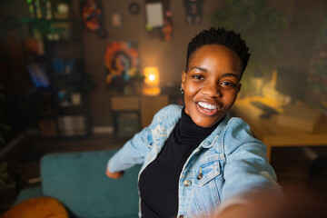 Cheerful african american woman takes selfie standing in the living room smiling wearing casual...