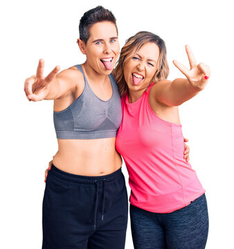 Couple of women wearing sportswear smiling with tongue out showing fingers of both hands doing victory sign. number two.