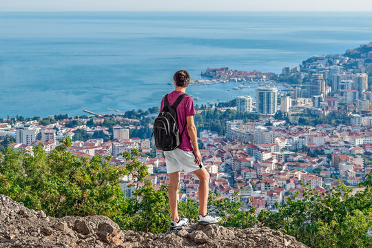 A man with a backpack and in summer clothes looks from the top of a mountain at the roofs of Budva and the Adriatic Sea in Montenegro. Solo hitchhiking in the Balkans - Powered by Adobe