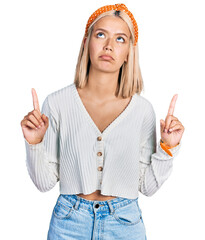 Beautiful young blonde woman wearing casual white sweater pointing up looking sad and upset,...
