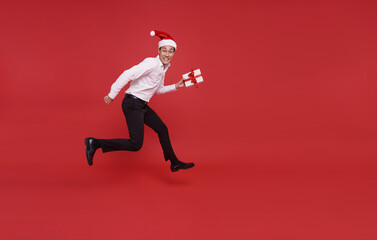 Fototapeta na wymiar Happy asian business man wearing santa hat holding pile of gift box and jumping celebration christmas day in office isolated on red background.