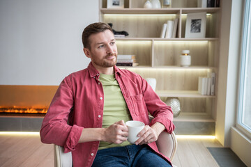 Positive pensive man drinking tea sitting in chair near fireplace looking at window at home....