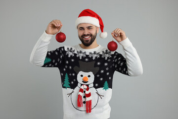 Happy young man in Christmas sweater and Santa hat holding festive baubles on grey background