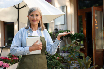 Happy business owner with tablet inviting to come into her flower shop outdoors, space for text