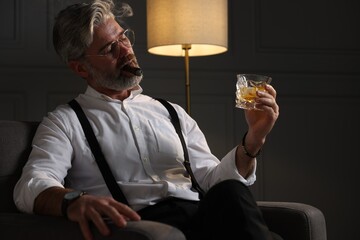 Bearded man with glass of whiskey smoking cigar in armchair indoors - obrazy, fototapety, plakaty