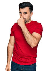 Hispanic man with beard wearing casual red t shirt smelling something stinky and disgusting, intolerable smell, holding breath with fingers on nose. bad smell