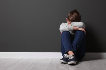Child abuse. Upset boy sitting on floor near gray wall indoors, space for text