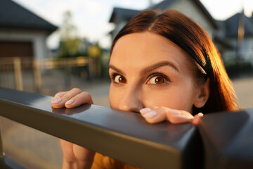 Concept of private life. Curious young woman spying on neighbours over fence outdoors, closeup