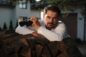 Concept of private life. Curious man with binoculars spying on neighbours over firewood outdoors