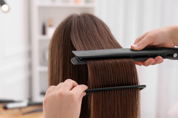 Hairdresser straightening woman's hair with flat iron in salon, closeup