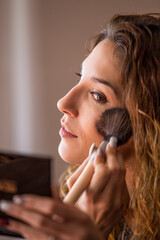 Mujer joven maquillándose echándose colorete en la mejilla mientras se mira en un espejo