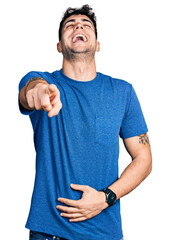 Young hispanic man wearing casual t shirt laughing at you, pointing finger to the camera with hand over body, shame expression