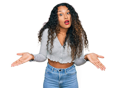 Young hispanic woman with curly hair wearing casual clothes clueless and confused expression with arms and hands raised. doubt concept.