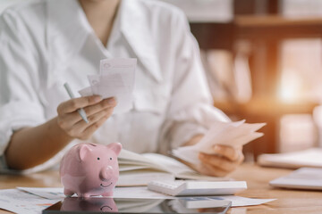 Woman sitting at desk managing expenses, calculating expenses, paying bills using laptop online, making household financial analysis, closer focus on the pink piggy bank. - obrazy, fototapety, plakaty