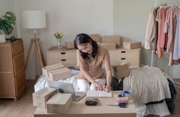 Busy Asian woman preparing package delivery box Shipping for shopping online taking note of order with laptop young start up small business owner online shop