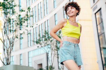 Young beautiful smiling hipster woman in trendy summer clothes. Carefree woman with curls hairstyle, posing in the street at sunny day. Positive model outdoors. Cheerful and happy