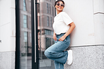 Young beautiful smiling hipster woman in trendy summer white t-shirt and jeans clothes. Carefree woman, posing in the street at sunny day. Positive model outdoors near wall. In sunglasses