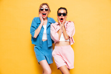 Two young beautiful smiling brunette hipster female in trendy summer clothes. Sexy carefree women near yellow wall in studio. Positive models having fun. Cheerful and happy. Shocked and surprised - obrazy, fototapety, plakaty