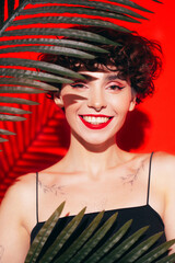 Fashion beauty portrait of young smiling brunette woman with evening stylish  makeup and perfect clean skin. Sexy model with curly short hair posing in studio. With red bright lips. Tropical palm leaf