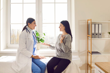Obraz premium Smiling female doctor talking with young woman while sitting on couch in medical office. Patient having consultation with GP, therapist during medical appointment in office. Health care and assistance