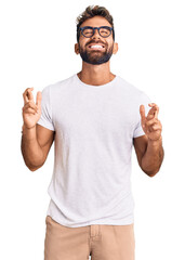 Naklejka premium Young hispanic man wearing casual clothes and glasses gesturing finger crossed smiling with hope and eyes closed. luck and superstitious concept.