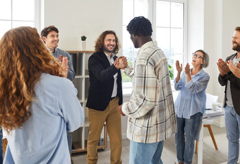 Obraz premium Diverse business team celebrating success and having fun in office. Two young men exchange handshakes while group of happy joyful mixed race colleagues clap hands and cheer. Teamwork success concept