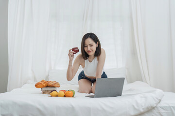 Obraz na płótnie Canvas Cute Asian woman sitting happily in bed with laptop and having fruit and morning snack.