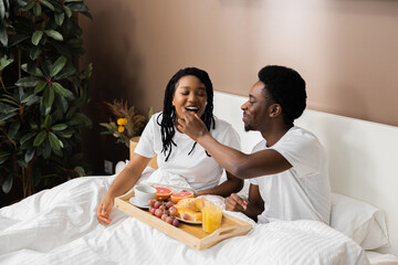 Beautiful African American couple in love sitting in bed drinking coffee and eating breakfast. Young man feeding dark skinned husband. Looking with love at each other.