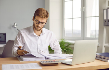 Portrait of young serious businessman in glasses working at home or at office. Man looking through paper financial documents with a pile of folders on his workplace analyzing company.