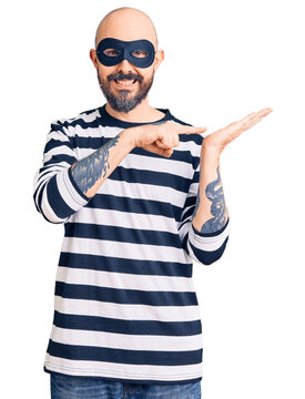 Young handsome man wearing burglar mask amazed and smiling to the camera while presenting with hand and pointing with finger.