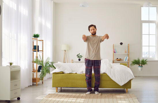 Sleepwalking. Somnambulist man gets out of bed in morning with closed eyes and outstretched arms. Young Caucasian man in pajamas walks like sleepwalker in bedroom. Sleep disorder concept.