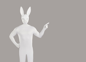 Man disguised in rabbit costume advertises something on blank grey copyspace background. Anonymous guy wearing white bodysuit and long eared playboy mask standing with hand on hip and pointing to side