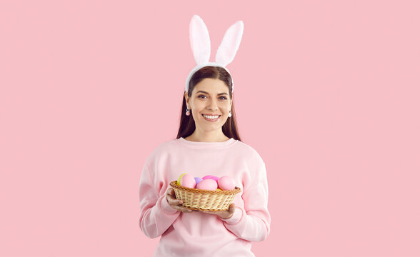 Portrait of happy brunette young woman with bunny ears holding basket with colorful eggs on pink background looking at camera. Celebrating easter, spring holidays, decorations for party concept.