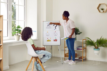 African American child girl sitting on chair and looking at school counselor or psychologist standing by white board, pointing at picture and asking questions. Education, children's psychology concept