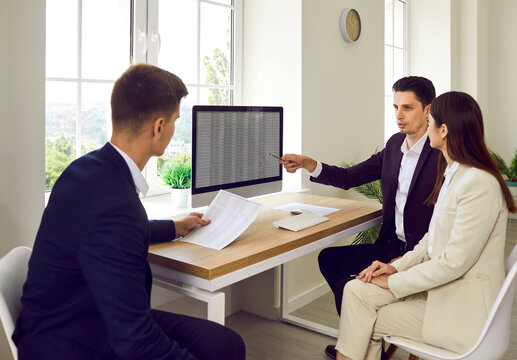 Business Team Looking At Accounting Data Sheet Together. Group Of People Sitting At Office Desk With Computer. Accountant Points At Screen, Shows Online Digital Spreadsheet And Explains Some Figures