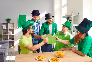 Happy friends celebrating St Patrick's Day. Group of Irish men in leprechaun hats having a party, sitting at table with chips and French fries, saying toast, clinking big mugs, and drinking green beer - obrazy, fototapety, plakaty