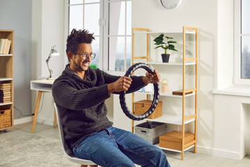 Side view photo of a young happy smiling african american guy keeping steering wheel and sitting on a chair at home. Man with a fashionable hairs style holding rudder of a car and driving.
