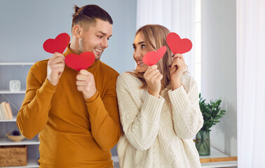 Young happy couple in love holding red paper hearts. Affectionate boyfriend and girlfriend wearing warm knitted jumpers standing in living room at home and looking at each other - Powered by Adobe