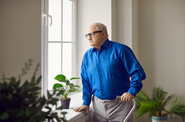 Portrait of pensive senior man looking at window. Lonely thoughtful elderly man with walking cane standing near window at home. Aging, loneliness, disability concept