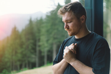 A man is praying near the beautiful panoramic window against the background of the forest. Christian man. Concept for faith, spirituality and religion.